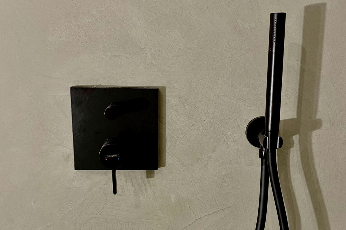 Moderne schwarze Duscharmatur an einer strukturierten, cremefarbenen Wand, neben einem Wandthermostat.